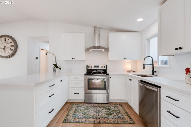 kitchen with light wood-style flooring, a peninsula, a sink, wall chimney range hood, and appliances with stainless steel finishes