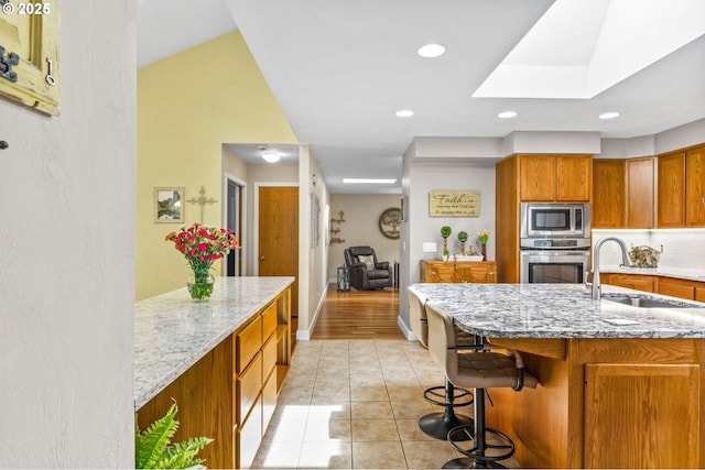 kitchen with light stone countertops, brown cabinets, a sink, a skylight, and stainless steel appliances