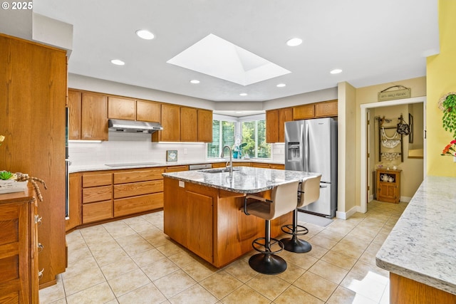 kitchen with under cabinet range hood, brown cabinets, stainless steel fridge with ice dispenser, backsplash, and a skylight