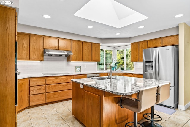 kitchen with stainless steel fridge, a sink, brown cabinetry, light tile patterned floors, and under cabinet range hood