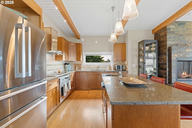 kitchen with appliances with stainless steel finishes, decorative light fixtures, sink, a kitchen island with sink, and wall chimney exhaust hood