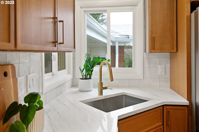 kitchen featuring decorative backsplash, brown cabinetry, light stone countertops, and a sink