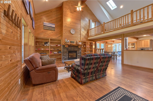 living room featuring high vaulted ceiling, ceiling fan, wooden walls, a fireplace, and a skylight