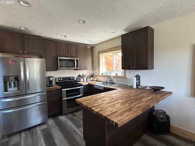kitchen with dark wood-style flooring, appliances with stainless steel finishes, a sink, a peninsula, and butcher block counters