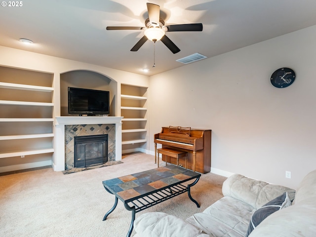 living room with a tiled fireplace, ceiling fan, built in shelves, and light colored carpet