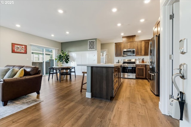 kitchen featuring a center island with sink, appliances with stainless steel finishes, open floor plan, wood finished floors, and light countertops