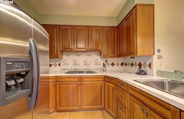 kitchen with a sink, cooktop, stainless steel fridge, brown cabinets, and backsplash