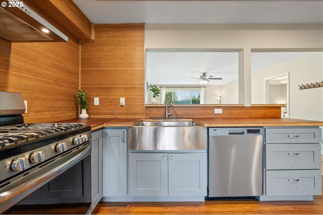 kitchen featuring stainless steel appliances, wall chimney range hood, ceiling fan, a sink, and light wood-type flooring