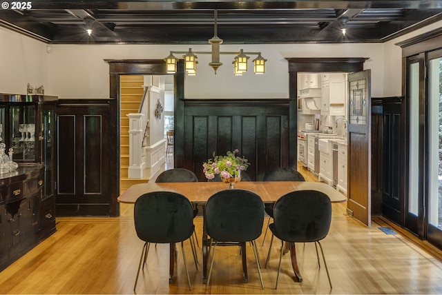 dining area featuring coffered ceiling, light wood finished floors, a wainscoted wall, beam ceiling, and a decorative wall