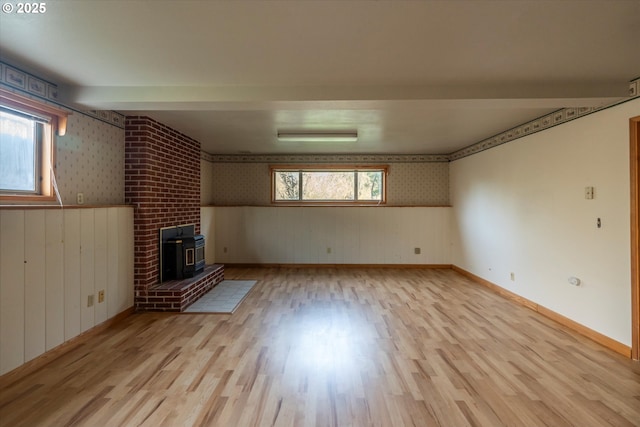 unfurnished living room with a wainscoted wall, baseboards, wallpapered walls, a wood stove, and light wood-type flooring