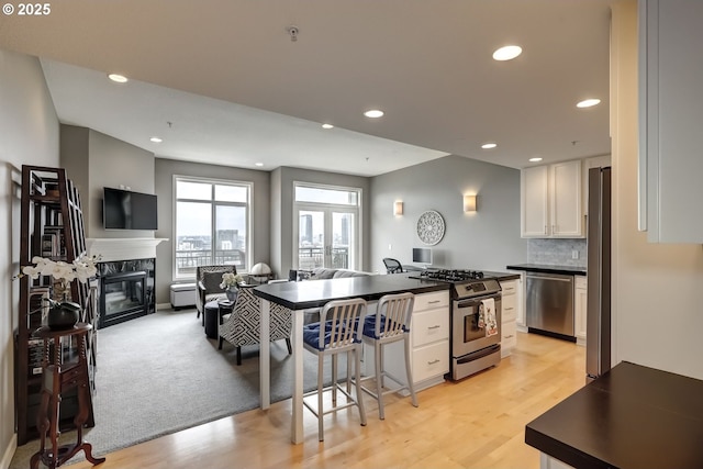 kitchen featuring a fireplace, stainless steel appliances, dark countertops, open floor plan, and white cabinets