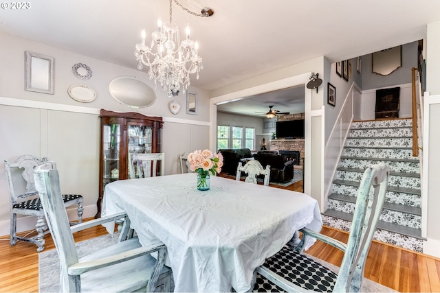 dining room featuring stairs, a stone fireplace, ceiling fan with notable chandelier, wood finished floors, and a decorative wall