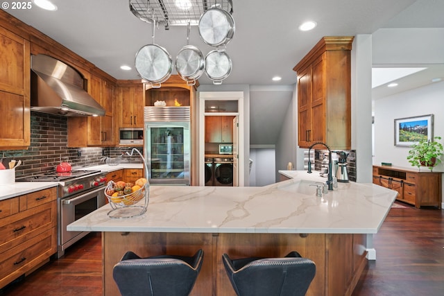 kitchen featuring built in appliances, wall chimney range hood, a sink, brown cabinets, and dark wood finished floors