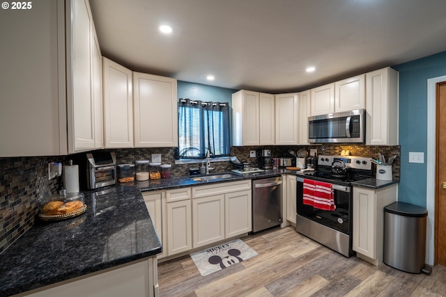 kitchen with stainless steel appliances, a sink, light wood-type flooring, backsplash, and dark stone countertops