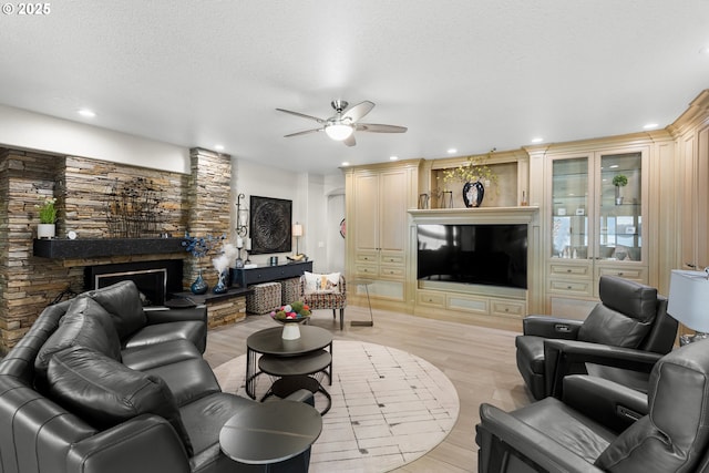 living room featuring ceiling fan, a stone fireplace, light hardwood / wood-style flooring, and a textured ceiling