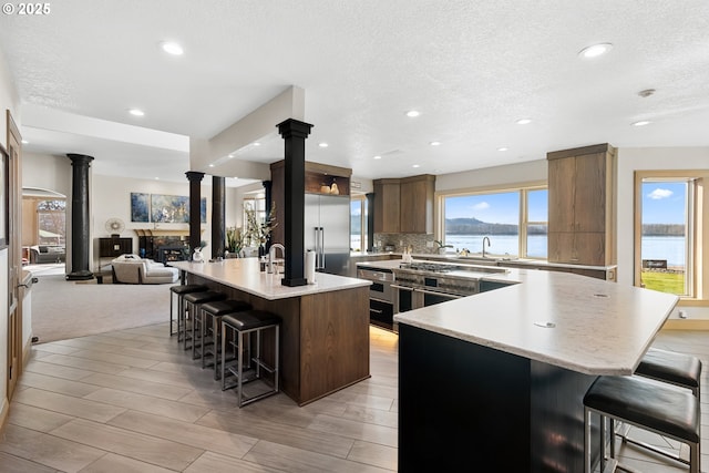 kitchen featuring a kitchen bar, ornate columns, a water view, stainless steel built in fridge, and a kitchen island with sink