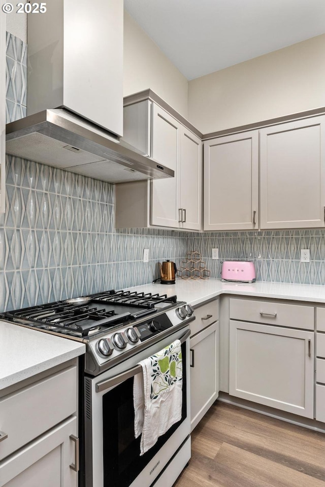 kitchen featuring stainless steel gas range oven, light wood finished floors, light countertops, wall chimney exhaust hood, and backsplash