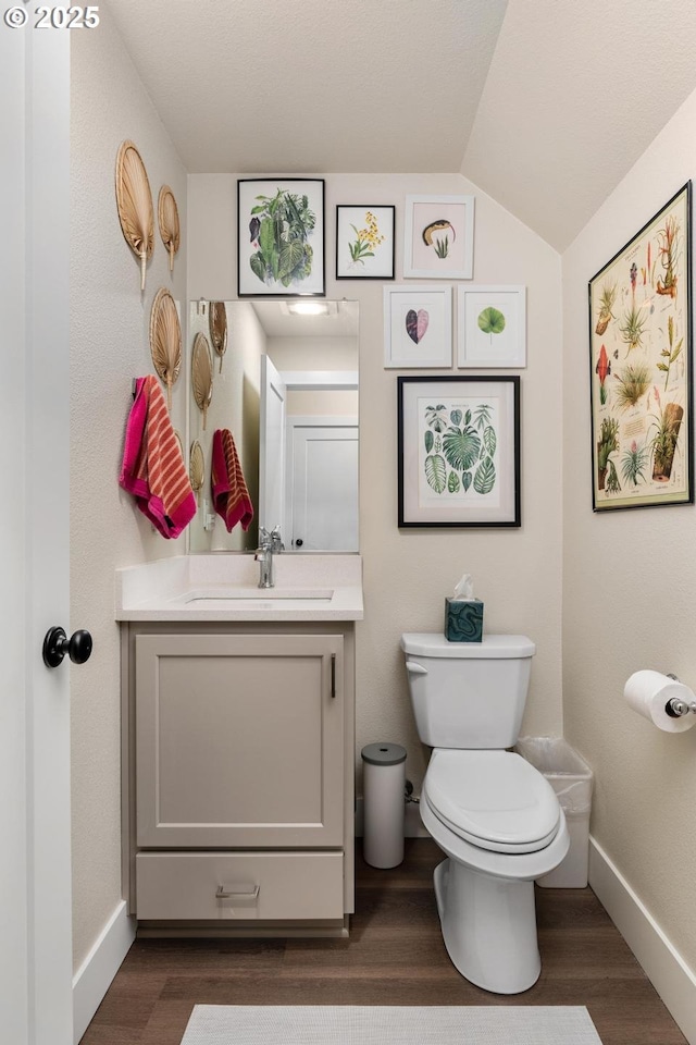 half bath with vanity, wood finished floors, toilet, baseboards, and vaulted ceiling