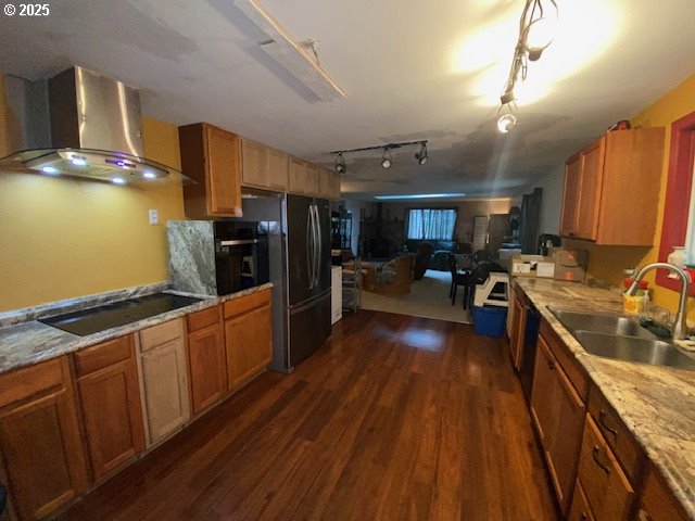 kitchen with brown cabinets, dark wood-style floors, black appliances, wall chimney exhaust hood, and a sink