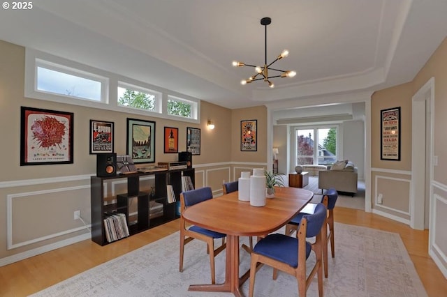 dining space with an inviting chandelier, wainscoting, a decorative wall, a raised ceiling, and light wood-style flooring