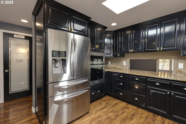kitchen with dark cabinets, appliances with stainless steel finishes, wood finished floors, and light stone countertops