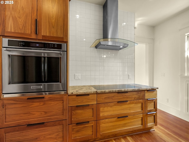kitchen with wall chimney exhaust hood, oven, black electric stovetop, tasteful backsplash, and light stone counters