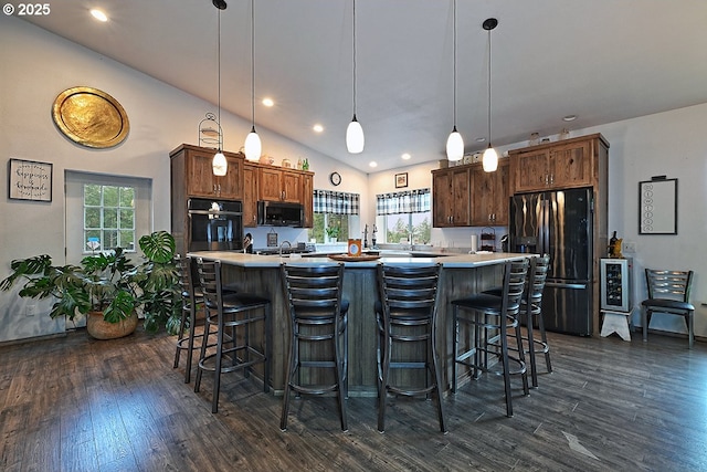 kitchen with dark wood-type flooring, hanging light fixtures, black fridge, a breakfast bar, and a center island with sink