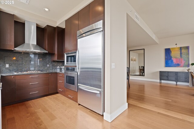 kitchen with built in appliances, wall chimney exhaust hood, light wood-style floors, backsplash, and recessed lighting