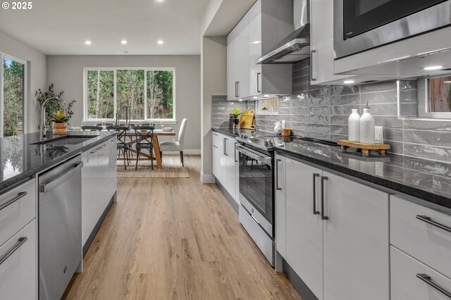 kitchen featuring a sink, appliances with stainless steel finishes, wall chimney exhaust hood, light wood-style floors, and white cabinetry
