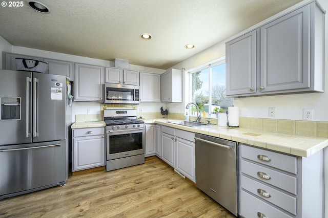 kitchen with light wood finished floors, tile countertops, gray cabinets, stainless steel appliances, and a sink