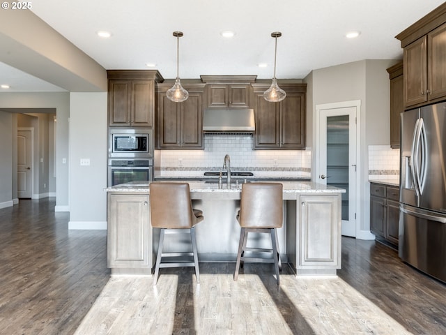 kitchen featuring light stone counters, decorative light fixtures, stainless steel appliances, and an island with sink