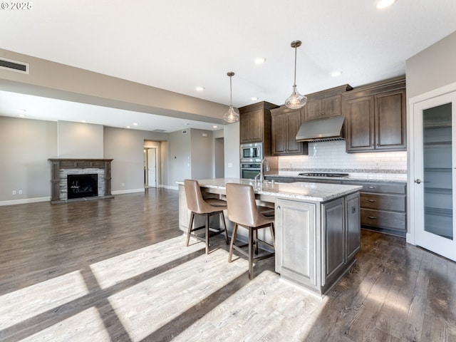 kitchen featuring tasteful backsplash, dark brown cabinets, hanging light fixtures, a center island with sink, and stainless steel appliances