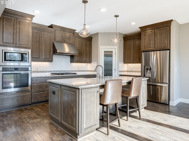 kitchen with a breakfast bar, an island with sink, hanging light fixtures, light stone counters, and stainless steel appliances