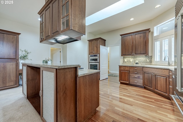 kitchen with double oven, a sink, decorative backsplash, light stone countertops, and light wood finished floors