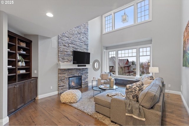 living area with baseboards, wood-type flooring, a stone fireplace, and a towering ceiling