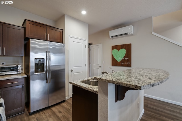 kitchen featuring a wall mounted AC, light stone counters, dark brown cabinets, stainless steel fridge with ice dispenser, and a center island with sink