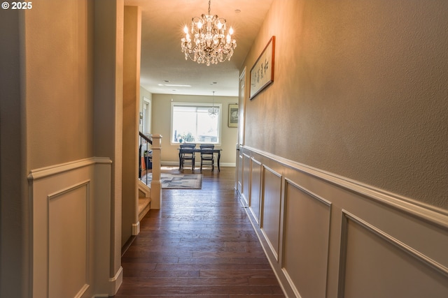 hallway with a wainscoted wall, stairway, dark wood-style flooring, a decorative wall, and a notable chandelier