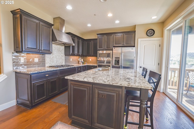 kitchen featuring stainless steel appliances, wood finished floors, a sink, wall chimney exhaust hood, and tasteful backsplash