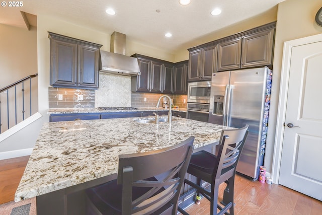 kitchen featuring light stone counters, wall chimney exhaust hood, appliances with stainless steel finishes, a sink, and a kitchen breakfast bar