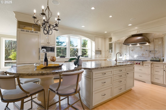 kitchen with tasteful backsplash, an island with sink, a kitchen breakfast bar, light stone counters, and wall chimney exhaust hood