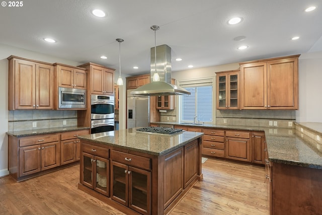 kitchen with built in appliances, island range hood, a kitchen island, brown cabinets, and glass insert cabinets