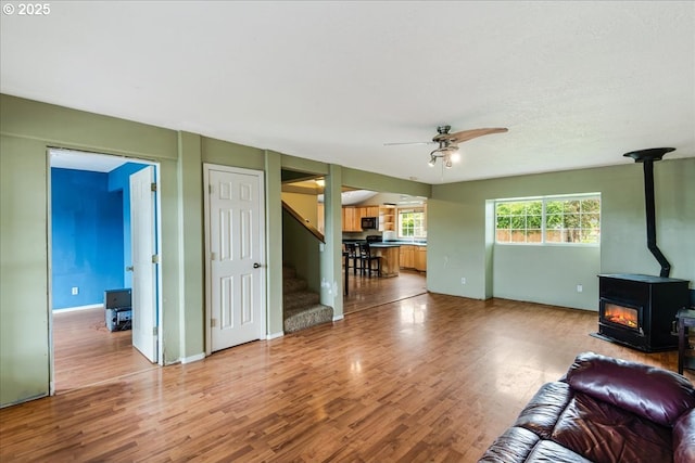 living area with a wood stove, ceiling fan, wood finished floors, stairs, and baseboards