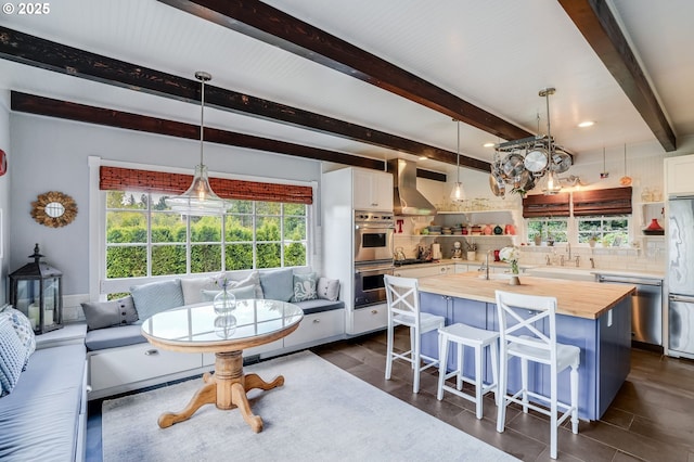 kitchen featuring butcher block countertops, stainless steel appliances, wall chimney exhaust hood, white cabinets, and beam ceiling