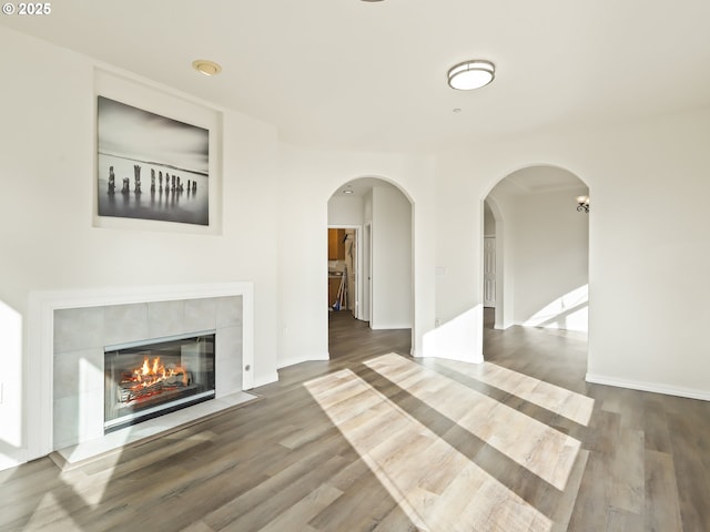 unfurnished living room featuring baseboards, a tiled fireplace, arched walkways, and wood finished floors