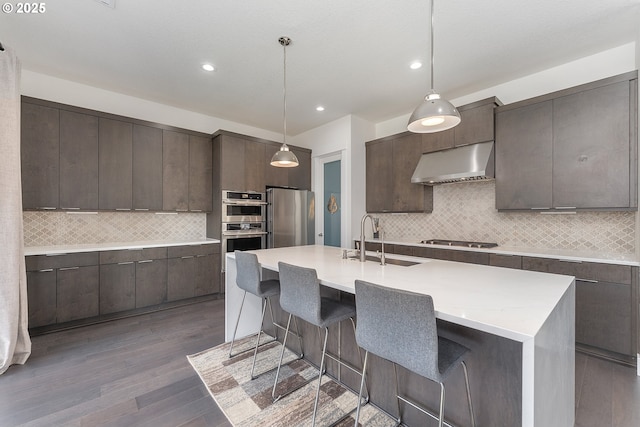 15875 NW Ridgeline St, Portland, OR 97229 - kitchen featuring wall chimney range hood, appliances with stainless steel finishes, dark brown cabinets, wood finished floors, and recessed lighting