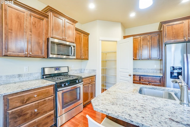 kitchen with appliances with stainless steel finishes, brown cabinets, light stone countertops, light wood-type flooring, and a sink