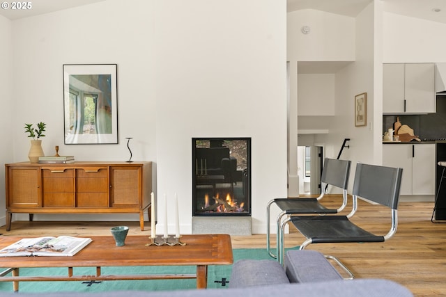 living room featuring light wood-style flooring, high vaulted ceiling, baseboards, and a glass covered fireplace