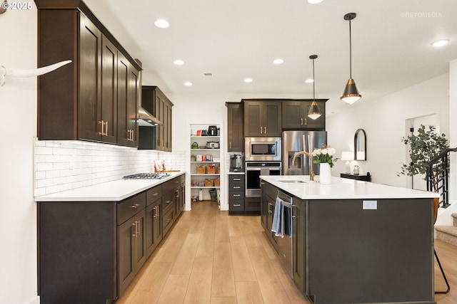 kitchen with a kitchen island with sink, stainless steel appliances, a sink, light countertops, and decorative light fixtures