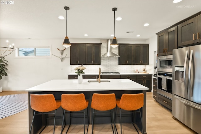 kitchen featuring a kitchen island with sink, stainless steel appliances, light countertops, wall chimney range hood, and a sink