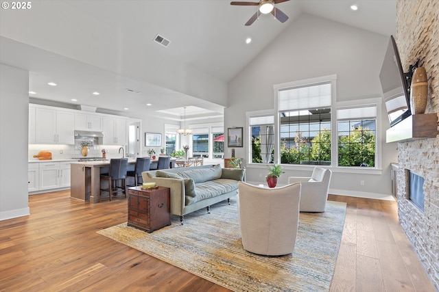 living room with visible vents, a ceiling fan, light wood-style flooring, a stone fireplace, and high vaulted ceiling